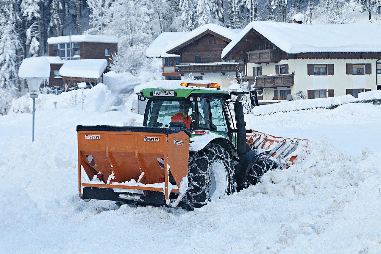 Traktor odśnieża zasypaną śniegiem drogę w górskiej scenerii. W tle ośnieżone domy i drzewa.