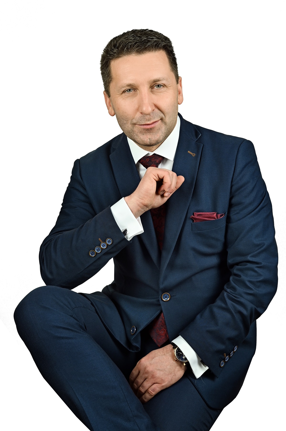 Portret mężczyzny w garniturze. Uśmiechnięty, elegancki, z zegarkiem. Siedzi na krześle, zrelaksowany. Tło białe.