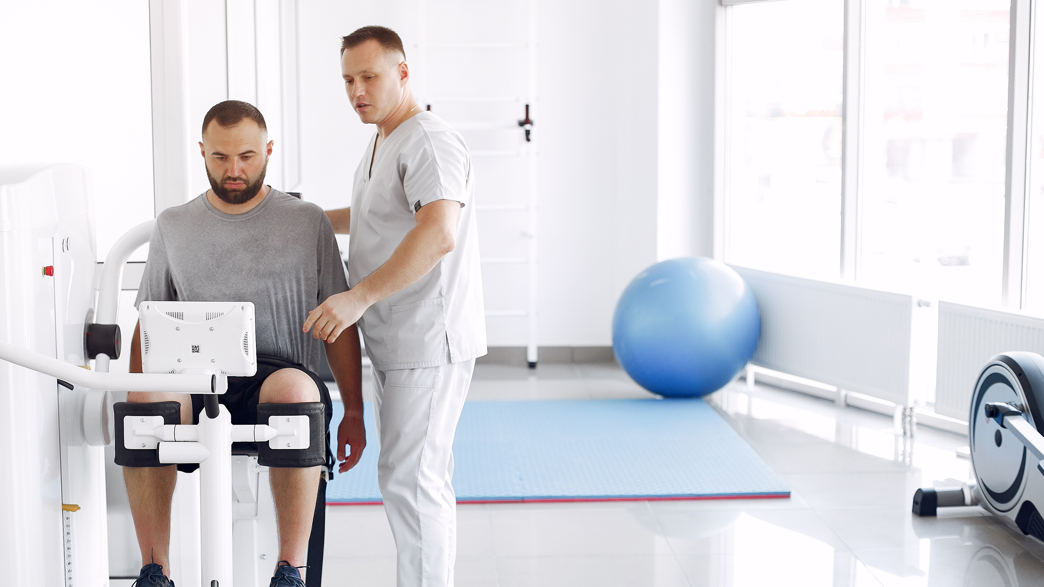 Fizjoterapeuta pomaga pacjentowi w rehabilitacji kolana na specjalistycznym sprzęcie w jasnym, nowoczesnym gabinecie.